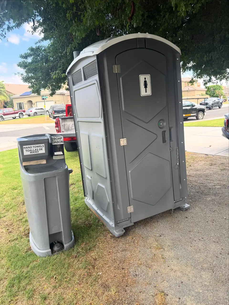 Grey standard porta potty beside a hand-wash station on a lawn